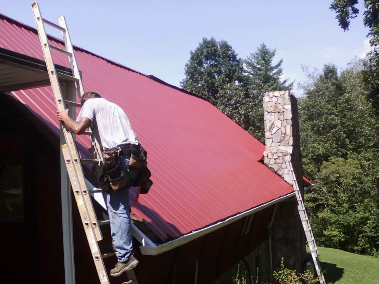 Roofer on a ladder beside a red metal roof and stone chimney — Soffit & Fascia Repair by Roofing Brothers in Mc Gaheysville